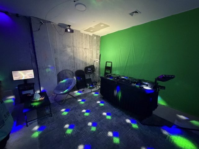 Blue and green light spots cover the floor around a DJ table, chairs and a green backdrop.