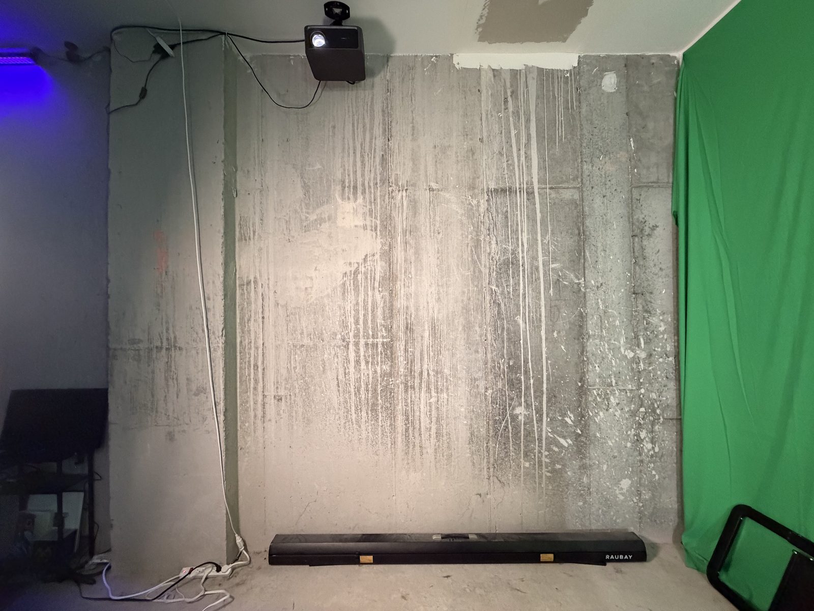 A bare concrete wall meets a green fabric backdrop, with a ceiling-mounted projector and a long case on the floor.