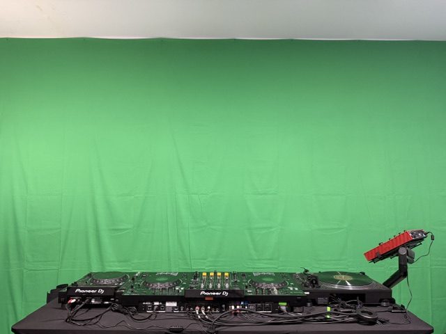 A black-covered DJ table holds decks and a mixer against a green fabric backdrop, with a red keyboard at one end.