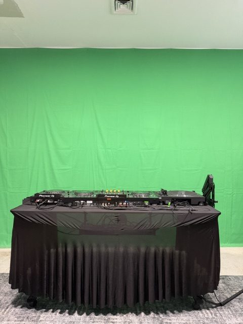 A green fabric backdrop spans the wall behind a black-draped table of DJ equipment.