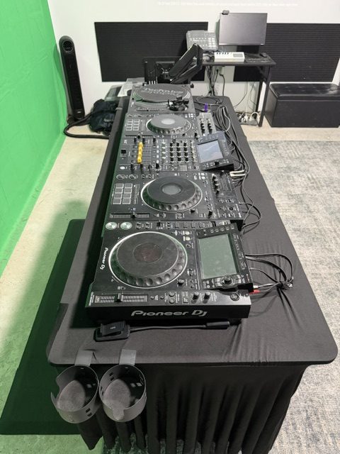 DJ decks and a mixer line a black-covered table, viewed from one end beside the green backdrop.