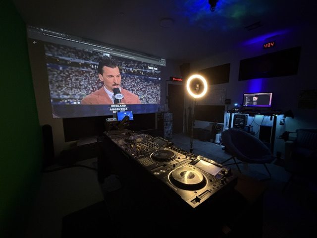 DJ decks face a projected sports broadcast, with a lit ring light and blue ceiling lighting beyond the booth.