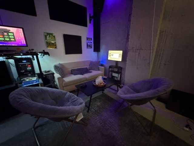Two round cushioned chairs face a low table beside a couch, with acoustic panels and purple lighting on the walls.